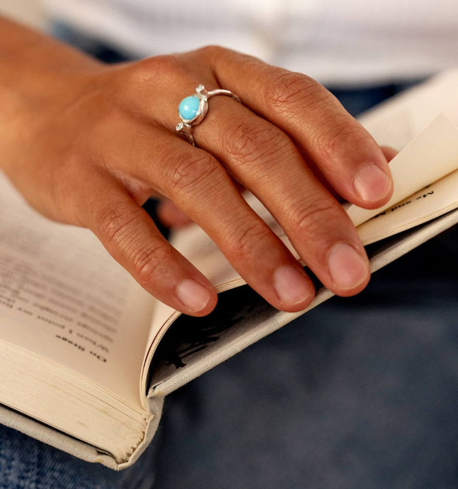 Hand holding an open book with a turquoise ring on a blurred background