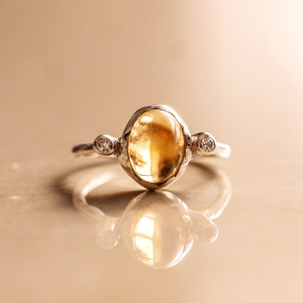 An organic style silver ring with a yellow citrine and two little diamonds on either side of the gemstone on a beige background.