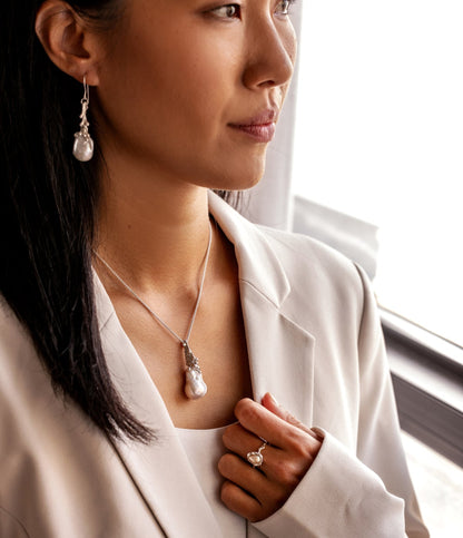 A woman wearing a pearl necklace, pearl earrings, and a pearl ring in front of a window.