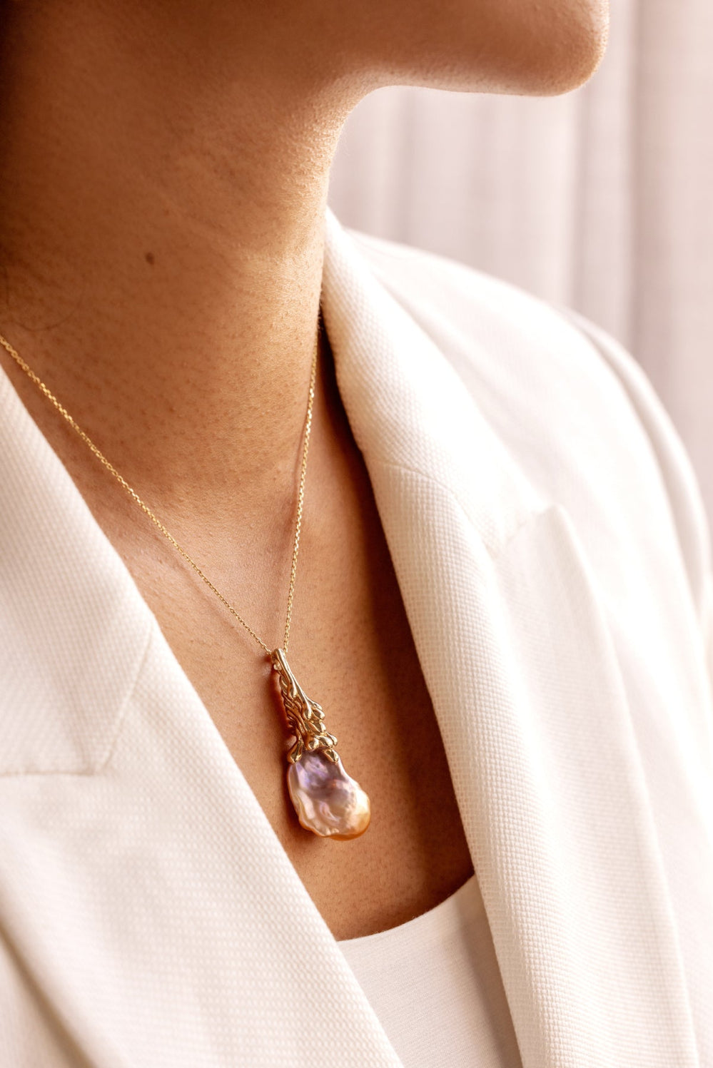 Gold necklace with an organic Baroque Pearl pendant worn by a person in a white outfit.