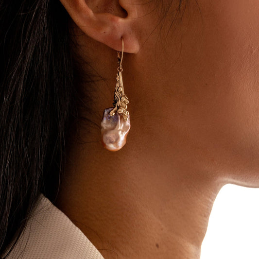 Close-up of a person wearing a gold earring with a pink organic baroque pearl pendant.